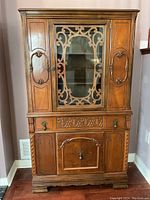 Front view of a vintage wooden display cabinet showing central glass door with decorative wooden grille, side wooden doors, a drawer with metal handles, and a lower wooden door with carved panels.