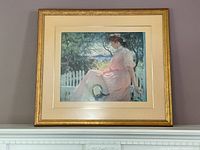 Full view of the framed art showing woman in pink dress sitting on a bench near fence, framed in a gold frame with cream matting.