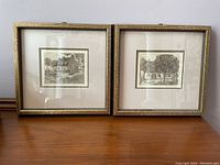 Two framed cottage scene art prints standing side by side on a wooden surface, against a pale wall.