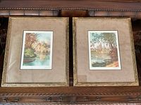Photo of two framed art prints side by side showing countryside water scenes with beige mats and gold-tone frames with wear.