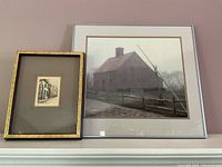 Two framed prints leaning against wall, one larger color rural house print, one smaller black and white street scene print with gold ornate frame.
