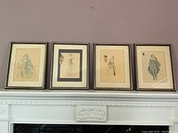 Four framed art prints showing costume design sketches displayed on a mantelpiece, in used condition with visible frame scratches and mat discoloration.