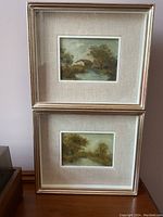 Pair of framed oil paintings stacked vertically, each with beige linen mat and silver frame, placed on a wooden surface.