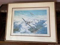 Framed art print showing two Spitfire planes flying above clouds with landscape below, signed at bottom.