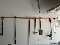 Wide view of garden tools hanging on wall mounted wooden strip showing full length of tools.