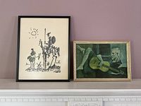 Two framed art prints displayed on a mantel against a purple wall: left is a black and white sketch of Don Quichote and Sancho Panza, right is a colored print of The Old Guitarist.