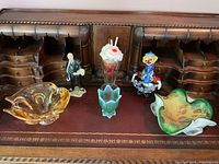 Includes all six items arranged on a wooden surface: two large decorative bowls, a blue votive holder, a sundae candle decoration, and two colorful glass figurines (clown and artist).