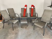 Wide view showing all five metal frame mesh patio chairs arranged in a semi circle on concrete floor. Two red camping chairs packed in fabric carry bags rest on two of the patio chairs.