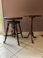 Two wooden furniture items on tiled floor against wall: an antique adjustable piano stool with round top and clawfoot casters, and a small pedestal side table with scalloped edge top and tripod clawfoot base.