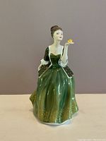 Front view of Royal Doulton figurine Fleur dressed in a green gown with gold designs holding a small yellow flower.