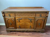 Front view of wooden sideboard showing three drawers and three cabinet doors with carved details and metal drop handles.