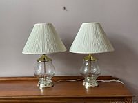 Pair of crystal table lamps with pleated white shades unlit on wooden surface against plain wall.