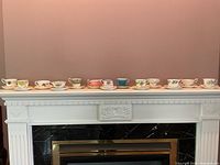 Photo of all 12 assorted vintage teacups and saucers displayed in a row on a white mantelpiece against a beige wall.