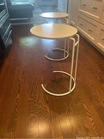Two white side tables with round tops and metal bases shown on wooden floor near kitchen cabinets and couch. Tables have C-shaped bases allowing them to slide close to furniture.
