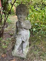 Front view of weathered concrete cherub statue standing in garden, showing detailed face and object held in hand.