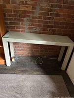 Front view of white melamine console table against a brick wall on a dark wooden floor with a portion of carpet visible.
