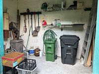 Wide view of shed interior showing green and black wheeled trash bins, garden hose coiled on wall, and multiple garden tools hanging including shovels, rake, and pitchfork.
