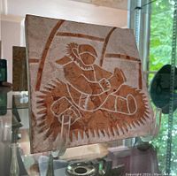 Front view of the square Inuit stone carving with clear acrylic stand showing the detailed figure and artist signature.