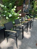 Four black mesh outdoor chairs with wooden armrests arranged on stone patio in front of garden foliage