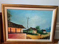 Full view of the framed painting showing a rural scene with houses, a dirt path, trees, and three human figures under a blue sky with birds.