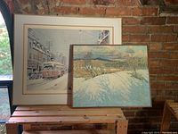 Two framed prints leaning against a brick wall; the canvas print of a snowy landscape (Tom Thomson) in front, and behind it a framed streetcar urban scene print with a white mat (Arto Yuzbasiyan)