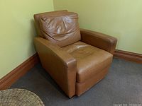 Tan genuine leather Roots armchair positioned in a corner against yellow walls and dark brown baseboards on carpeted floor, showing angled view of right side and front.