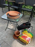 Photo showing square glass top metal folding table with potting soil, black metal dust pan, terra cotta pots and black bag with garden rocks on patio floor