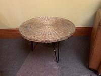 Side view of round wicker top side table with metal hairpin legs placed on carpeted floor next to couch.