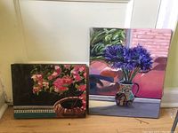 Photo showing both oil paintings side-by-side leaning against a wall, clearly showing size difference and floral subject matter.