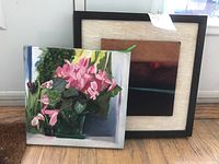 Photo showing the 10x10 canvas painting of pink cyclamen flowers leaning against two framed artworks, one larger with dark wood mat and fabric, and another smaller abstract piece.