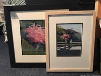 Two framed and matted floral artworks, one black-framed with a pink flowering tree, one natural wood frame with pink flowers in a vase.