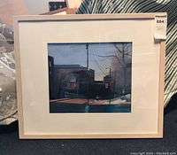 Front view of framed and matted original painting showing urban street scene with buildings, traffic light and car at dusk with light wood frame.