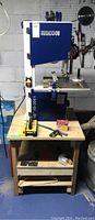 Full view of blue Rikon band saw mounted on wooden stand with clamp and blade box
