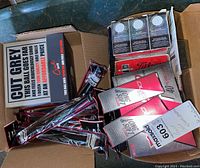 Overhead view of open cardboard box containing multiple packaged SuperStroke Cross Comfort grips, boxes of golf balls (including Titleist, Maxfli, Cut Blue), and a pack of Tread cleats.