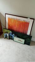 Five framed wall art pieces leaning against a wall: large orange/red abstract tree painting, dark green abstract painting, small evergreen tree print, horizontal vintage landscape with windmill, and blue flower painting close together.