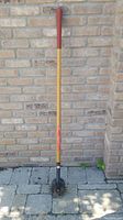 Single garden edger standing upright against brick wall outside, showing full length with red handle grip and metal blade head at the bottom.