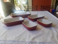 Five red ceramic Kitchenaid baking dishes arranged on a white cloth. Includes one large lasagna dish and four smaller rectangular baking dishes with handles.
