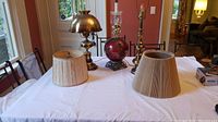 Full view of three lamps on table with two lampshades. Brass and red decorative lamp identifiable. Both lampshades shown, one smaller pleated shade, one larger beige shade.