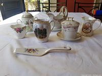 The entire lot arranged on a white sheet showing six different ceramic pieces of varying sizes, including a lidded sugar bowl, small pitchers, a creamer, a lidded bowl and saucer, and a figure jug.