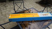 Blue and yellow Senior Telemaster model plane box with the model name and illustration of the plane on top, shown placed on an outdoor glass table.