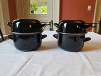Two black enamel mussel pots with lids on a dining table, showing the front and side views of both pots stacked with lids fitted.