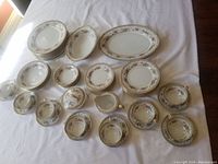 Full collection of Noritake Sommerset china dishes laid out on a white cloth, showing cups, saucers, plates, bowls, and serving dishes with floral and gold trim pattern.