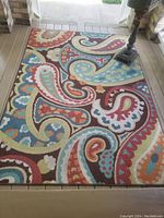 Top view of large colorful indoor/outdoor mat laid on porch, showing paisley and floral pattern in multiple colors.