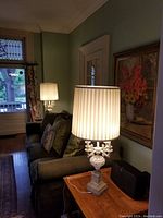 Photo showing one table lamp illuminated on a wooden side table with a green wall and a painting behind it, highlighting the pleated white shade and ornate base.