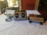 All three appliances displayed: Breville glass kettle, Black and Decker 4-slice toaster, Hamilton Beach panini press on table.