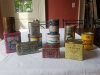 Full view of all 12 vintage tin cans displayed on a white cloth on a table with a red background and window light
