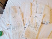 Three sets of white or cream towels, some with floral decoration, and two cream bath mats on a white sheet.
