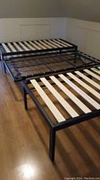 Two black metal twin bed frames with wooden slats set on a wood floor. The frame in front is a standard twin, the one behind has a trundle underneath that slides out for additional sleeping or storage.