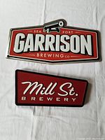 Top view of the Garrison Brewing Co. metal sign and Mill Street Brewery wooden sign laid flat on a white surface.