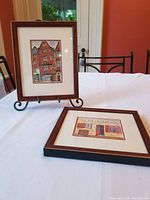 Two original framed watercolours placed on white table with iron chair and red wall background, showing timber-framed house and a storefront painting.